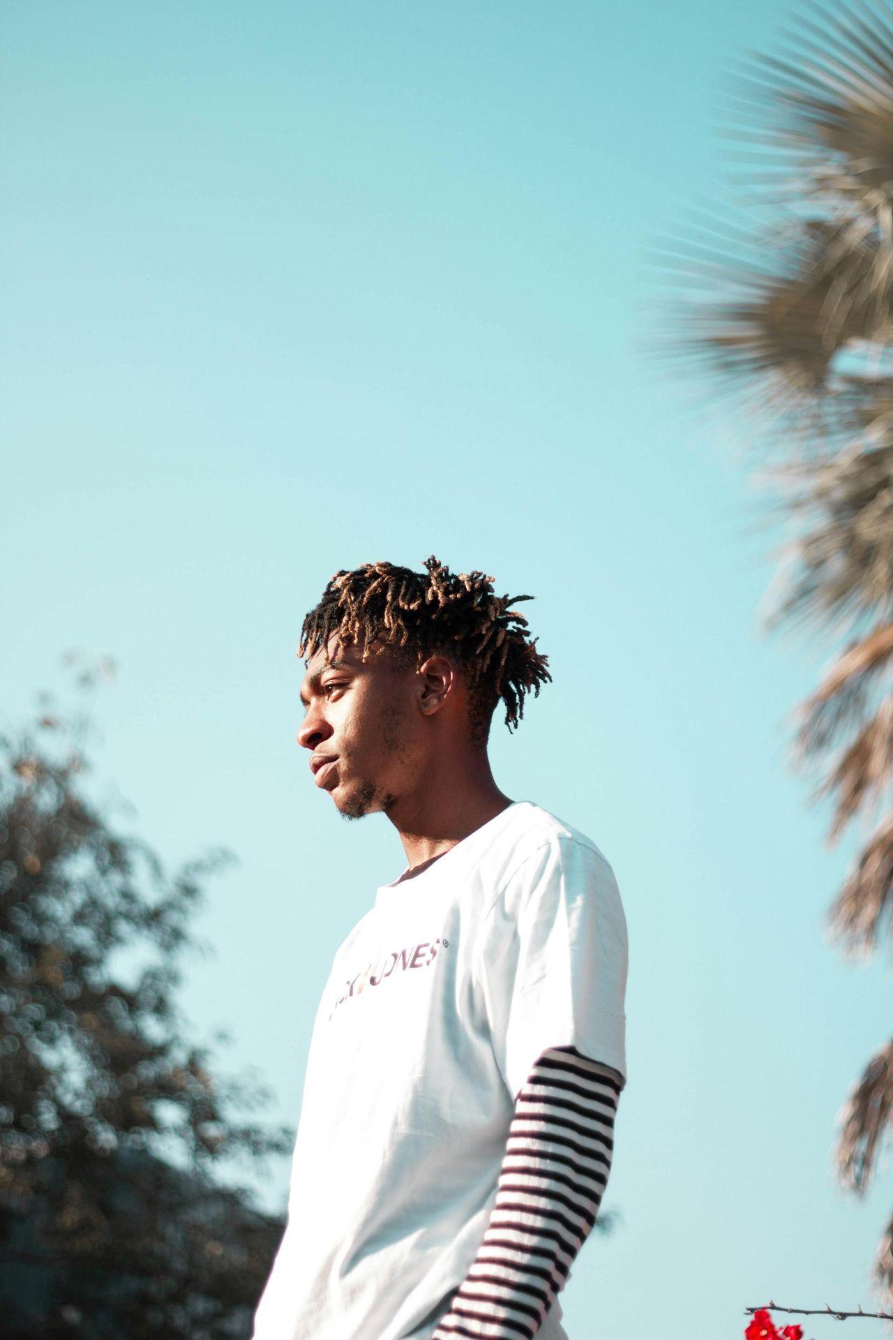 Person with dreadlocks wearing a white and striped shirt, standing outdoors against a clear blue sky.