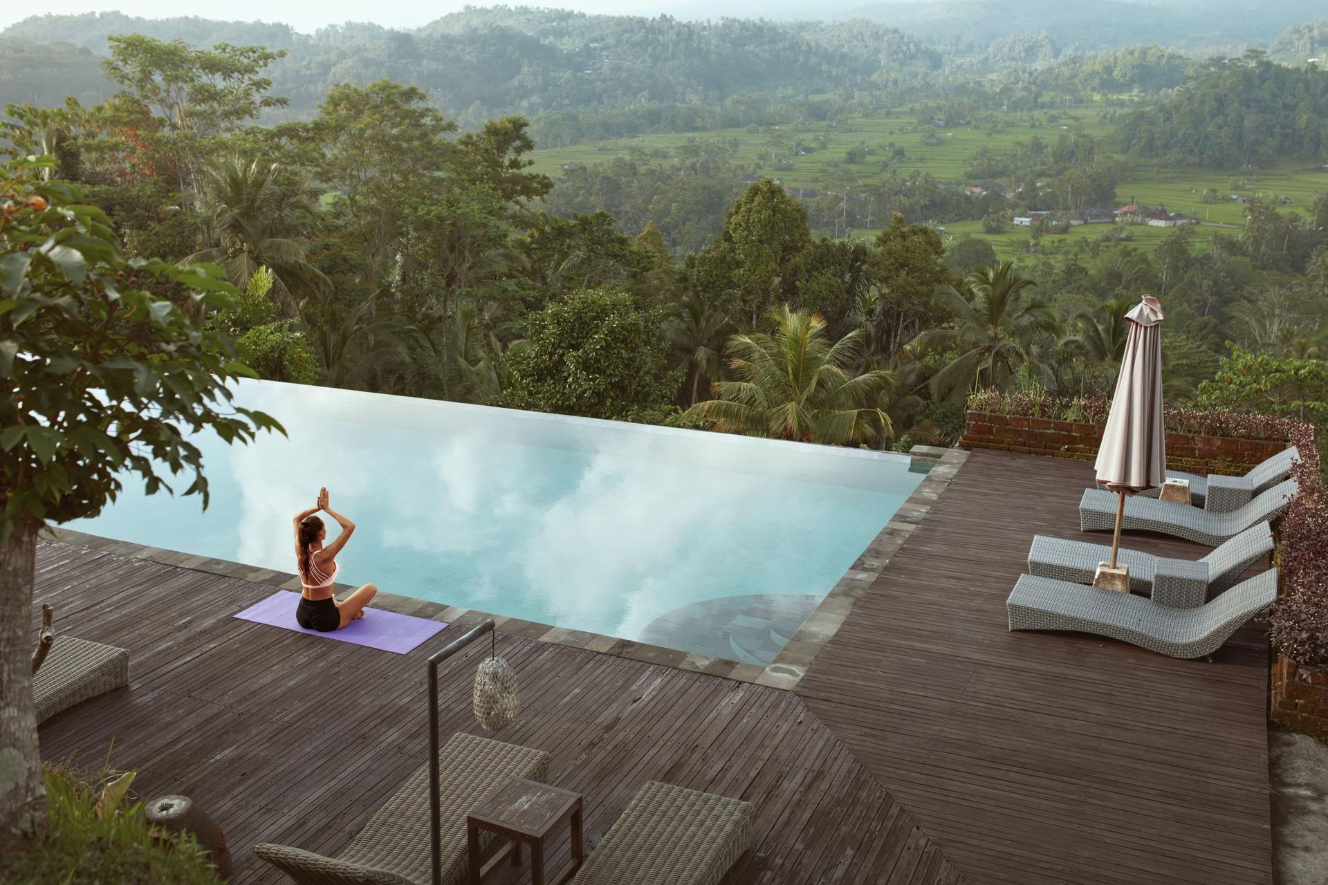 Beautiful Girl Doing Yoga On Poolside In Morning In Bali, Indonesia. Young Slim Woman In Sportswear Sitting In Lotus Pose Or Padmasana On Sport Mat Near Infinity Pool Against Tropical Landscape.