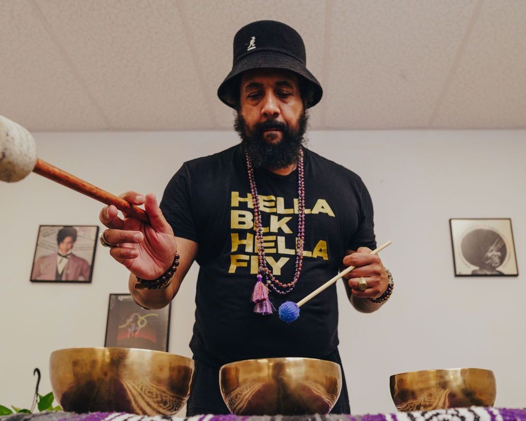 Person wearing a hat and beaded necklace, playing bronze singing bowls with a mallet.