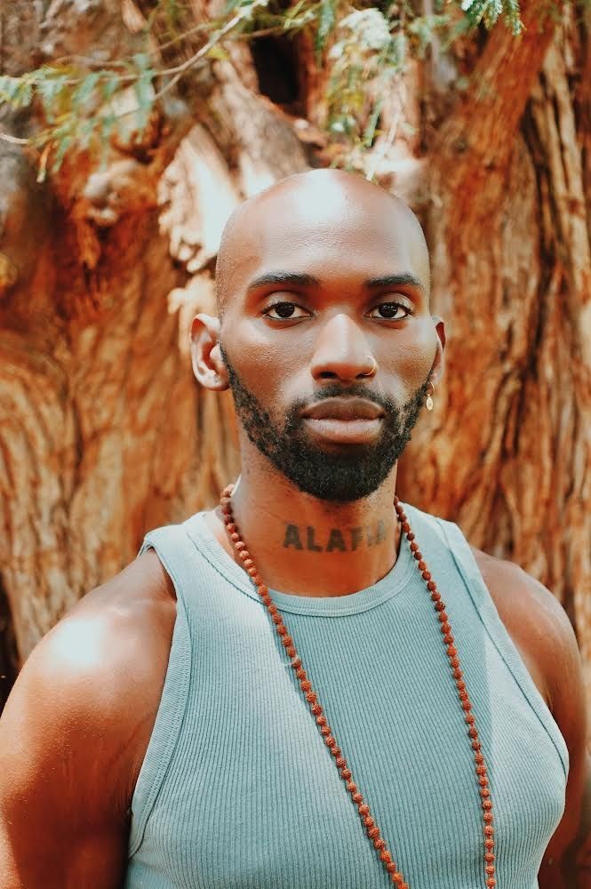 Person in a blue tank top wearing a beaded necklace, standing in front of a tree.