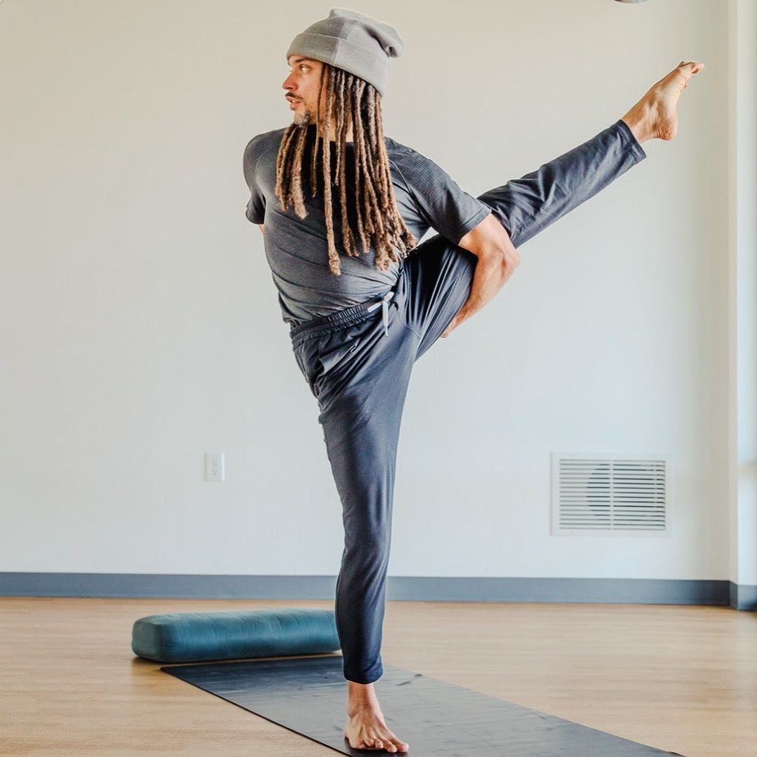 Person in gray clothing performing a high leg stretch on a yoga mat in a bright room.