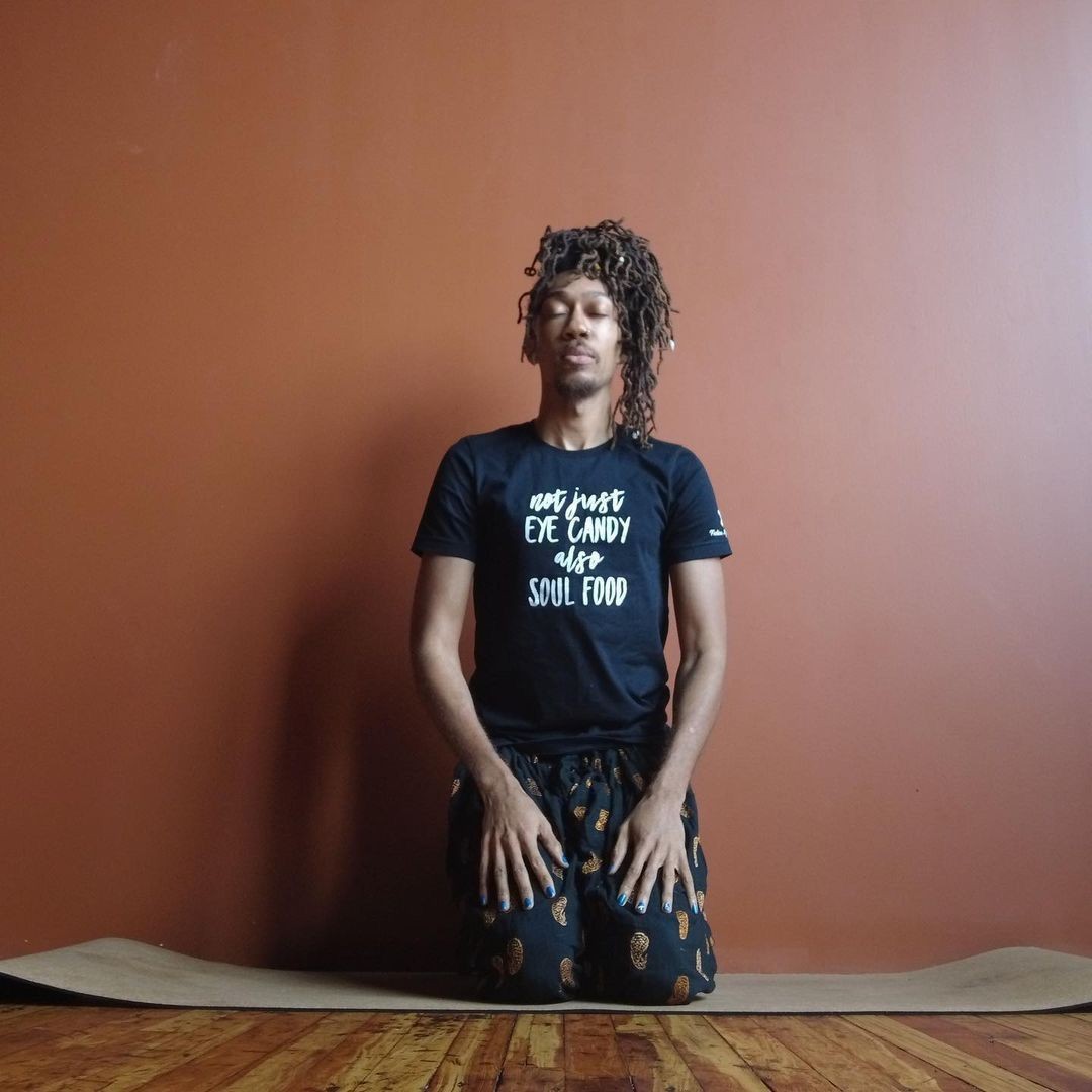Person in a black shirt with text, kneeling on a yoga mat against a brown wall.