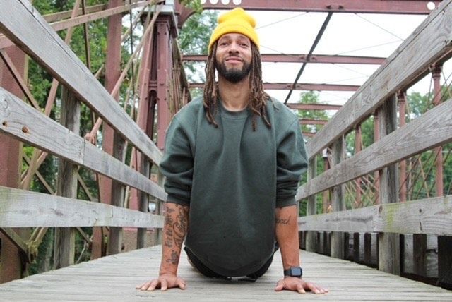 Man with a yellow beanie and green sweatshirt doing a yoga pose on a wooden bridge.